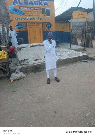 This image depicts a street scene in Panshekara, Nigeria, captured in the late afternoon or early evening, at 6:29 PM on January 26, 2026. The sky appears overcast, creating soft, diffused lighting.
The main subject is a man standing towards the right foreground, dressed in a white long-sleeved kaftan, white trousers, and dark flip-flops. He wears glasses and poses with his right hand on his chin, looking at the camera with a slight smile. The ground beneath him and extending into the foreground is dusty and uneven dirt.
Behind him, to the left, another man in a similar white garment stands beside a wooden stall displaying bright yellow fruits, possibly oranges. A child in a red shirt is seated on a low stool near this stall. Between the central figure and the fruit stall is a weathered, rusty white chest cooler or freezer. A large sack filled with yellow items is placed on the ground near the stall.
The background features a vibrant yellow and blue building with various texts and illustrations painted on its facade. The prominent text reads "AL BARKA Accessories Ventures... Dealers in all types of handset new phone Bluetooth, screen guard cover, power bank Mpx. car mpa. chargers. usa, batteries, etc." Below this, in what appears to be a local language, is "MUNA BADA SARIN KATI ANAN" (likely "We sell recharge cards here"), followed by two phone numbers: "0706984540.07083690563". Logos for "MTN" and "glo" are also visible on the building. The building has a yellow door. To the right of the building, a separate "MTN" sign is mounted on a pole. The scene suggests a local commercial or market area.