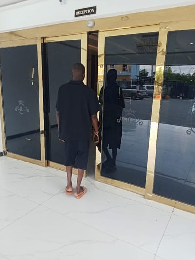 A black male adult, wearing a black shirt and black shorts, stands in front of an automatic sliding glass door with gold trim. The door is labeled "Reception" in gold lettering. The man is looking down at the floor. There is a second individual, whose face is not visible, reflected in the glass. The individual is wearing dark clothing. The floor is tiled in white. There is a reflection of a car in the glass doors. This scene is most likely located at a hotel or office building in Ujevwu, Nigeria. The coordinates (latitude: 5.472, longitude: 5.817) are in the city of Ujevwu, which is located in the Delta State of Nigeria.