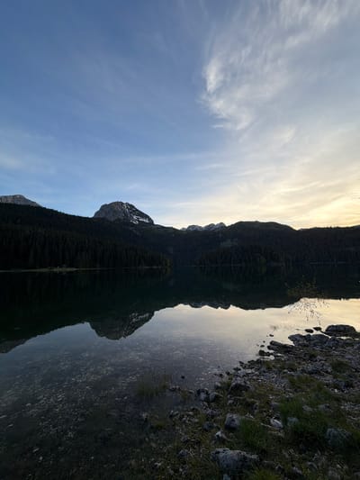 The image depicts a landscape scene, mirrored vertically. The upper half shows a sky with wispy clouds and a blue hue transitioning to lighter tones near the center. The lower half appears to be a reflection in water.
In the center, a dark, elongated mass stretches vertically, resembling a mountain range covered in trees. There's a clear line dividing the mountain and its reflection. On the right side, the land meets the water with rocks and grass. The mirrored reflection of these rocks and grass gives texture to the reflection. This scene appears to be a view of the Durmitor National Park, located in Pitomine, Montenegro, known for its mountains and lakes. There are no people visible in the image.