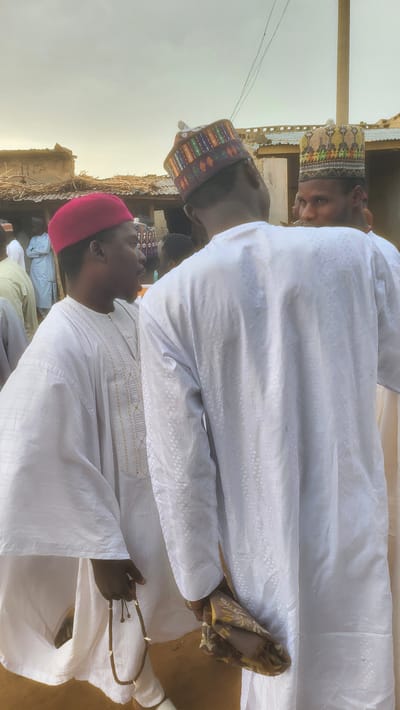 The image, rotated 90 degrees clockwise, captures a gathering of at least three men outdoors in Jogana, Nigeria. When viewed upright, the scene shows the men standing on what appears to be sandy or dirt ground, against a backdrop of simple, weathered structures, possibly with corrugated iron roofs or mud walls, indicating a rural or semi-urban setting. Thin utility wires are visible against a bright, hazy sky, suggesting daylight, likely early morning or late afternoon due to the soft, diffused lighting.

In the foreground on the left, a man is dressed in a white traditional robe with subtle embroidery and a vibrant pink/red embroidered cap. He holds a string of prayer beads (tasbih) in his left hand, looking downwards or towards the person beside him. Behind him, slightly to the right, another man in a similar white robe wears a darker, multi-colored patterned cap and is looking in the direction of the first man. On the far right, a third man's head and upper body are visible, also in a white robe and a lighter, patterned cap. He looks directly at the camera with a neutral expression.

All men wear traditional long white robes and embroidered caps, which are common attire in the region. The presence of prayer beads suggests a religious or spiritual context for the gathering, or simply personal devotion. The overall atmosphere appears calm and communal. There is no visible text in the image.