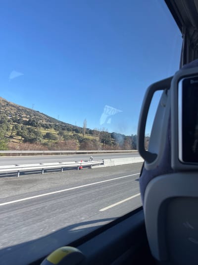 This image captures a bright, sunny day scene from the perspective of a vehicle, likely a bus or car, traveling along a highway. The camera is angled, showing a broad expanse of clear blue sky dominating the upper left portion of the frame.
To the right of the sky, a landscape of rolling hills or mountains is visible, densely covered with green trees and some bare deciduous trees. Utility poles or power lines can be seen stretching across the hillside. In the middle distance, on the hillside near the road, a small, light plume of smoke or dust is visible, suggesting a minor fire, burning, or some form of activity.
The lower right part of the image features a multi-lane highway or expressway. A concrete barrier separates the main lanes from a narrower service road or shoulder. Guardrails run alongside this inner road. Parked on this service road is a white vehicle, possibly a small truck or van, with a bright orange cone placed nearby, indicating a stop or roadside work.
Inside the vehicle, parts of the window frame and interior fittings are visible at the top and right edges. A handle or armrest is prominent at the top center. A distinct reflection of reversed text, appearing to read "OZOE" or "30Z0", is visible on the window, likely originating from an object within the vehicle.
The scene depicts travel through a rural or semi-rural area under clear, bright daylight. Given the location context of La Losa, Spain, the landscape of tree-covered hills alongside a modern highway is consistent with the region. The image conveys a sense of a journey on a clear day.