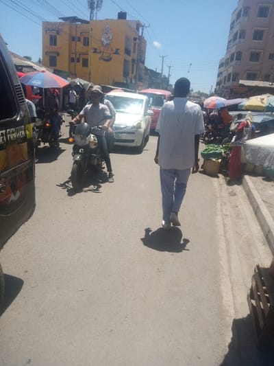 This image captures a street scene in Mtopanga ward, Kenya, under clear, sunny conditions. The perspective is from behind a pedestrian walking away from the camera.
In the immediate foreground on the left, the rear side of a dark-colored vehicle, possibly a van or minibus, is partially visible, displaying "STREET FIGHTER" graphics. On the right, a paved asphalt road extends into the background.
A male individual is walking away from the camera, occupying the right side of the road. He wears a white short-sleeved shirt, light blue pants, and white shoes, with his shadow visible directly behind him on the pavement.
To the left of this pedestrian, a dark-colored motorcycle with a prominent round headlight is being operated by a male wearing a light-colored t-shirt and a wide-brimmed hat. Another individual is seated as a passenger behind the driver. This motorcycle is positioned ahead of a stationary white sedan, which bears a license plate ending in "L". Behind the white sedan, a red van or minibus is partially visible.
Along the right side of the street, multiple market stalls are established, shaded by assorted colorful umbrellas (red, blue, and yellow). Sacks or baskets containing green vegetables are visible on the ground beside these stalls. A concrete curb and drainage channel separate the market area from the road.
In the background on the left, a multi-story yellow building prominently displays "Never Say Never Say" and "SUPA LOAF" in large text, alongside a baker's logo. Utility poles with overhead wires traverse the sky. Further down the street on the right, another multi-story building with a light brown or pinkish facade is visible. Numerous other pedestrians and motorcycles are interspersed throughout the background, contributing to the busy environment.