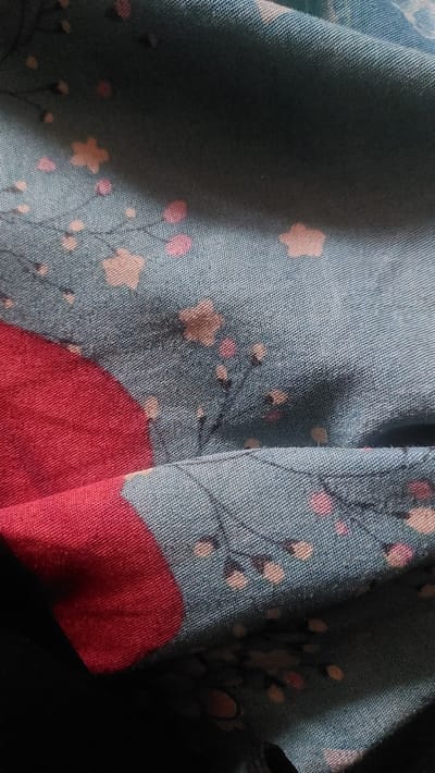 The image shows a close-up view of a teal fabric with a floral pattern. The fabric is creased, suggesting it is draped or folded. The pattern consists of delicate branches with small pink and light brown flowers, and some darker circular elements. A large, irregular patch of bright red is visible in the lower left portion of the image, partially obscuring the patterned fabric. The texture of the fabric is visible, showing a woven material. The lighting creates shadows and highlights on the folds of the fabric. There are no discernible people, specific actions, or a recognizable environment related to New Nyanya, Nigeria. The image is solely focused on the fabric and its pattern.