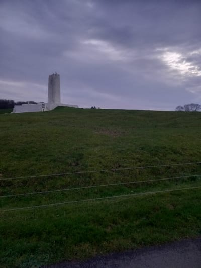 The image shows a large monument with a tower set against a cloudy sky. The monument is on a field of green grass. Several power lines are to the right of the field along a road. Trees are at the top of the field and at the bottom of the monument.