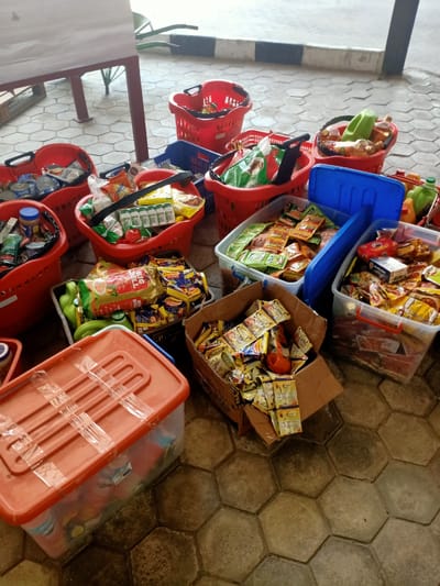 The image shows several red plastic shopping baskets and a few blue and orange plastic storage containers filled with various food items and household supplies. The items include packets of seasoning, noodles, cooking oil, canned goods, and other groceries. The containers are arranged on a floor with hexagonal tiles, appearing to be in a storage area or a distribution point. Based on the prompt, the location is Dongo, Nigeria, although there are no specific landmarks or identifiers in the image to confirm this. There are no people visible in the frame.