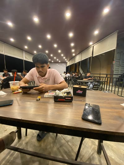 The image captures a casual nighttime scene in what appears to be an indoor or semi-outdoor cafe or restaurant in Keudeublang, Indonesia. The photograph is taken from a slightly elevated perspective, looking down at a wooden table and its occupants, and is rotated on its side.
In the foreground, a young man with dark hair, wearing a light pink polo shirt, is seated at a dark wooden table. He is intensely focused on a black smartphone he holds with both hands. To his right, on the table, is a small yellow lighter. Closer to him is a black box with "Aura" written in white cursive, and another black box with "BOLD NEW WORLD" visible on its side, along with the partially visible word "BATT". A plastic cup with a drink and a red straw is positioned further down the table to his left, next to another lighter and a phone.
The setting is illuminated by numerous circular white lights embedded in a dark brown or reddish-brown ceiling, creating a warm glow. In the background, other individuals, mostly men, are visible seated at tables, some also looking at their phones or socializing. A black metal railing separates the interior space from an exterior area where parts of a building and a motorcycle are faintly visible. The floor is concrete, and parts of chair legs are seen beneath the table.
The main activity is people relaxing and engaging with their mobile devices in an evening social setting. The presence of food packaging and drinks suggests a dining or refreshment establishment.