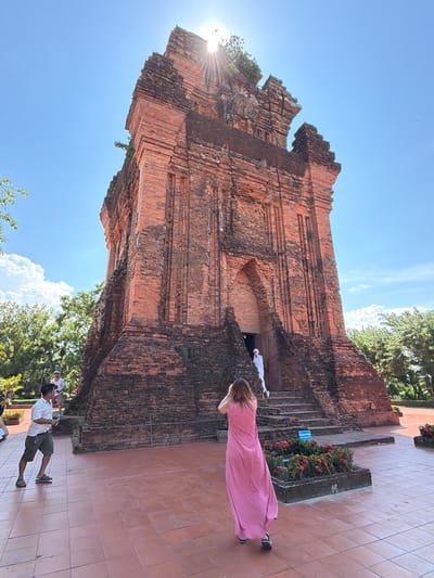 This is a vertically oriented, eye-level, medium shot of the Po Nagar Cham Towers in Tuy Hoa Ward, Vietnam. The ancient brick towers, a notable landmark, dominate the frame, with their weathered red brickwork and intricate layered architecture. The sun is shining brightly, casting a lens flare through the top left of the image, suggesting it is daytime. The sky is a clear, vibrant blue with a few wispy white clouds. Lush green trees surround the towers, adding to the natural setting.

In the foreground, on the right side of the image, a person wearing a long, flowing pink dress stands on a tiled walkway, facing the towers. Their back is to the camera. To the left of the towers, on a set of stairs, another person wearing white clothing appears to be walking or standing. Further down the walkway, another person in a white shirt and olive green shorts is seen from the side, also facing the towers. The overall mood is peaceful and tourist-like, with individuals exploring a historical site. There is a blue sign visible on a planter box near the stairs, but the text is illegible. The scene appears to be set outdoors in a warm, sunny climate.