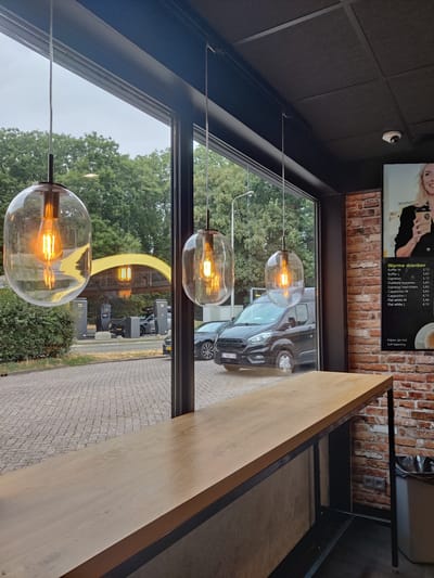 This is a medium-shot, eye-level photograph taken from inside a building looking out through a large window. The interior features a wooden counter with a dark metal frame. Three large, clear glass pendant lights with visible Edison-style bulbs hang in front of the window. The window looks out onto an outdoor scene with trees, a paved area, and what appears to be a fuel station with electric vehicle charging stations. A dark-colored van is parked in front of one of the charging stations, and a black car is parked behind it. The exterior wall of the building is visible on the right, made of red brick, with a poster displaying a menu of "Warme dranken" (warm drinks) and prices. A small white security camera is mounted on the dark ceiling. The overall lighting suggests it is daytime, possibly overcast. The location is identified as Eijsden, Netherlands, at a fuel station with EV charging. There are no people clearly visible in the scene.