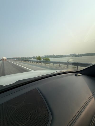 An outdoor, slightly hazy daytime, eye-level, wide-angle shot captures a view from inside a vehicle driving on a highway. The highway stretches forward, with a white line dividing two lanes on the left. In the distance, a white tanker truck is visible in the left lane. A metal guardrail runs along the right side of the highway, separating it from a body of water. On the far bank of the water, a city skyline with buildings and trees is discernible. The sky is a pale, hazy blue, suggesting it might be early morning or late afternoon, or that there's dust or smoke in the air. The interior of the car is partially visible in the foreground, including the dashboard and a speaker grille on the left. The scene appears to be on a bridge or elevated road, given the expanse of the water and the distant cityscape. The location is likely on the Ob River in Berdsk, Russia, as the cityscape and river are consistent with this geographical area.