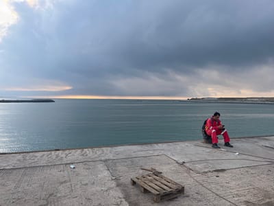 A man in a red jumpsuit sits on a bollard at the edge of a concrete pier, looking at his phone. Behind him, the water stretches to the horizon under a dramatic, cloudy sky with a hint of light breaking through. A wooden pallet lies on the pier in the foreground, and a distant landmass is visible on the right. The scene is overcast, with a calm sea.