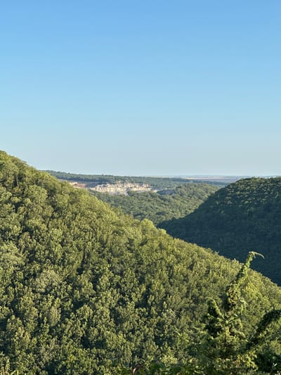 This is a landscape photograph taken on a clear, sunny day. The image depicts rolling hills covered in dense green foliage, characteristic of a forest. In the distance, a lighter colored cliff face is visible, suggesting a quarry or a natural rock formation. The sky is a clear, pale blue, dominating the upper portion of the image. There are no people or animals visible in the frame. The scene appears to be a natural environment with evidence of human activity (the quarry). The location is the city of Pryvorottia Druhe, Ukraine.