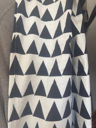 A close-up shot features a fabric with a bold geometric pattern. Alternating rows of white and dark-colored triangles create a dynamic, visually striking design. The triangles appear uniform in shape and size, arranged in an alternating, repetitive manner. The fabric is draped or folded, creating subtle shadows and highlights that emphasize the texture and dimension of the pattern. The bottom part is laying on a gray cloth.