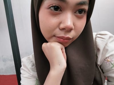A young woman, wearing a dark brown hijab and a light-colored shirt with floral embroidery, is captured in a close-up, selfie-style portrait. Her right hand is resting on her chin, with her left arm visible. The background consists of a white wall and a dark vertical element on the left side, possibly a door frame or furniture. A sliver of red carpet is visible in the lower left corner. The lighting is soft, highlighting her facial features and the details of her clothing. The image appears to have been taken indoors.