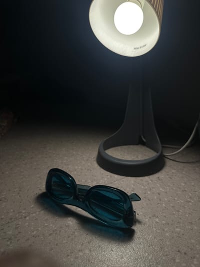 A desk lamp illuminates a pair of blue sunglasses resting on a speckled grey surface. The lamp is partially visible, with a white lampshade emitting a bright light. The sunglasses are positioned to the right of the lamp's base. The background is dark.
The city of Le Pré-Saint-Gervais, France is not visible in the image.