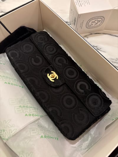 A black Chanel clutch with a repeating "C" and circle pattern and a gold clasp is nestled in a white box with tissue paper. A black dust bag is visible next to the clutch. To the left of the box is a white Diptyque candle labeled "BAIES" with a weight of 190g. The tissue paper has "BREAK ARCHIVE" printed in green. The box is lined with black trim.