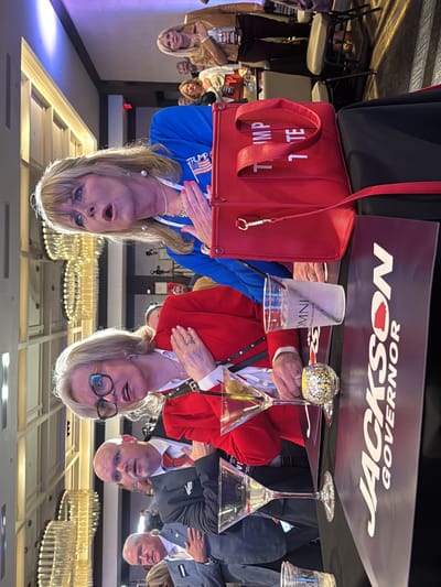 The image shows a group of people standing at attention, with their right hands over their hearts, presumably during an event. The people are seated at tables with beverages and campaign signs for "Jackson Governor". One woman is wearing a blue blazer with a "Trump" pin and has a large red tote bag with "Trump" on it. The room is brightly lit with chandeliers. The drink cups show "Omni Atlanta Hotel at CNN Center". Therefore, the location is the city of Atlanta, United States.