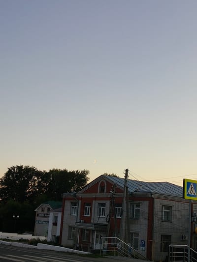In Сеченово, Russia, a twilight scene unfolds. A crescent moon is visible in the pale sky above a two-story building constructed of white brick with red trim. The building appears to be a commercial establishment, with signage indicating "Аптека" (Pharmacy) and "ЗОЛОТО ПОДАР" (Gold Gift) on one of its facades. A pedestrian crossing sign, a blue square with a white stylized human figure, stands to the right of the building. The foreground shows a road with a painted pedestrian crossing. Trees line the left side of the image. The overall impression is one of a quiet evening in a provincial town.