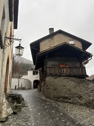 The image presents a narrow, wet cobblestone street located in Visp, Switzerland, under an overcast sky with visible light precipitation. The scene is devoid of people.

To the immediate left, a multi-story building with a light-colored facade, detailed with reddish-brown trim around arched recesses, features an ornate black wrought-iron lantern affixed to its upper section. A short set of irregular stone steps descends from this building to the street level.

Dominating the right side of the street is a multi-tiered building complex. Its base consists of a rugged stone retaining wall. Above this, a traditional dark wooden chalet-style structure with a prominent dark-tiled, pitched roof and rain gutters extends, featuring a covered balcony with a carved wooden railing. Small yellow flowers are visible on the balcony. Higher up, a lighter, possibly stucco-finished building with its own pitched roof and a central chimney stack rises, displaying a single rectangular window in its upper section.

Further down the street, on the left, a white building with a dark arched entrance and a small window fitted with dark shutters is visible. A curved white wall extends from this structure, along which a black streetlamp and a small stone fountain or trough are positioned.

The cobblestone street appears dark and reflective due to moisture. In the distant background, snow-dusted mountains are visible beneath the uniformly pale, gray sky.
