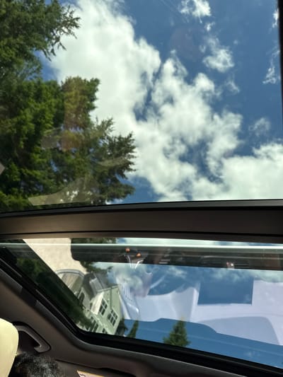 The image captures a view through a car's sunroof on a bright, sunny day. The sky is a vibrant blue, dotted with fluffy white clouds. Lush green trees are visible, their branches reflecting on the sunroof's glass. In the reflection, we can see a light green, two-story house with multiple windows, along with a street and other cars. The interior of the car is partially visible, showing a beige headrest and a person's dark, curly hair in the lower left corner. The scene appears to be in a residential area. The specific location in Issaquah, United States of America, cannot be determined from the visual information alone.