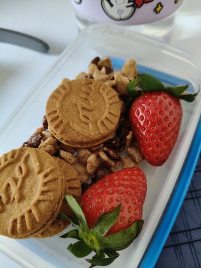 The image displays a clear plastic food container filled with snack items, viewed from a slightly elevated, top-down perspective. Inside the container are two whole, ripe red strawberries, each retaining its green calyx. Alongside them are two round, light brown sandwich biscuits featuring an embossed decorative pattern. A loose mixture of light brown cereal flakes and darker clusters, consistent with granola or breakfast cereal, forms the base beneath and around the strawberries and biscuits. The container has a visible blue rim on its right edge. In the background, a portion of a light-colored cylindrical drinking vessel, possibly a water bottle, adorned with a cartoon-like character in light purple and white tones, is visible at the top. A curved, dark grey object is partially seen in the upper left background. The items rest on a light-colored, flat surface. The specific location within Riccione, Italy, is not identifiable from the visual information provided. No individuals are present, and no specific actions or interactions are depicted.
