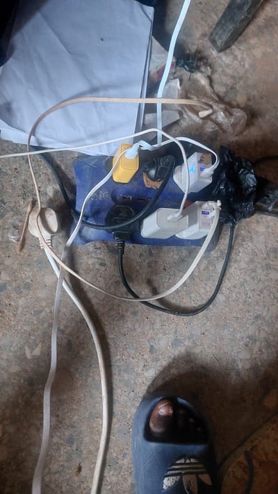 The image shows a close-up view of a power strip overloaded with multiple adapters and cables. The power strip is blue and sits on a rough, concrete-like surface, possibly a floor. Among the connected items are at least two white USB charging adapters, one with blue accents, and a yellow adapter. Several white and black cables are tangled around the power strip. To the left of the power strip, a single-prong plug is visible. In the lower right corner, a portion of a dark blue sandal with white stripes is visible, with a bare foot inside, indicating the presence of a person. The background is cluttered with what appears to be discarded plastic bags and paper. The scene suggests a somewhat disorganized environment, likely indoors, given the indoor appliances and the presence of a foot in a sandal. The location is specified as New Nyanya, Nigeria.