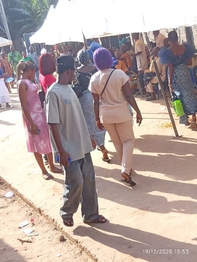 The image depicts an outdoor social gathering in Isinyi-Nando, Nigeria, captured on 19/12/2025 at 15:53. The ground is a light brown, dusty or sandy dirt path with scattered debris including paper and small objects. In the midground, multiple white canopies provide shade, under which numerous individuals are either standing or seated on chairs.
In the foreground, a male individual is central, facing largely away from the camera with his head turned slightly to his right. He wears a light grey short-sleeved t-shirt with dark text (possibly "TIA"), baggy light blue jeans, and dark sandals. He has a dark baseball cap worn backward and holds a blue object, possibly a mobile phone, in his left hand. To his immediate left and slightly behind him stands a female with light-colored braided hair pulled back. She wears a sleeveless pink dress with a tie-dye pattern and light-colored flat sandals, also holding a blue object in her left hand.
Further back and to the right of the central male, a female with vibrant purple hair is walking away from the camera. She wears a beige short-sleeved top and matching beige trousers, with a dark bag slung over her left shoulder. Another female, partially obscured behind her, wears a black and white patterned dress or skirt with a dark head covering. On the far right, a woman in a dark patterned dress with prominent ruffled sleeves is visible, carrying a bright green object or bag.
The background shows more attendees under canopies, dressed in a variety of attire including patterned fabrics. Green foliage from trees is visible in the upper left background. The scene is brightly lit by natural sunlight, casting distinct shadows on the ground, indicating an afternoon setting.