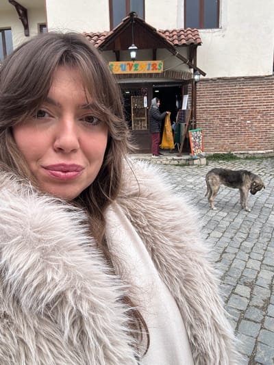 The image depicts a female subject in the foreground taking a selfie in Sighnaghi, Georgia. She is visible from the chest up, with light brown hair and bangs, light complexion, and light pink lipstick. She wears a shaggy, light beige or off-white faux fur coat over a lighter-colored garment. Her expression is a subtle smile as she looks directly at the camera.
In the midground, a "SOUVENIRS" shop is visible behind the subject. The shop entrance is framed in dark wood, with a colorful sign reading "SOUVENIRS" hanging above it. The building features a white plastered upper facade, a red-tiled roof, and two dark wall-mounted lanterns flanking the entrance. Inside the shop doorway, a person wearing a dark jacket and red trousers is observed holding up a yellow patterned textile item. To the right of the shop entrance, an outdoor display stand holds multiple small, colorful items.
To the right of the shop, a medium-sized dog stands on all four legs on the cobblestone pavement. The dog has multi-toned dark and light grey-brown fur and a yellow ear tag visible on its right ear. Its head is lowered towards the ground. The entire visible ground surface is paved with grey cobblestones. The lighting is consistent with daylight, possibly overcast.