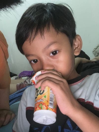 A young boy, likely of Asian descent, is captured in a close-up selfie, holding a cup of juice to his lips. His dark, tousled hair frames his face, and his dark eyes gaze directly at the camera. He is wearing a light-colored t-shirt with dark accents and a red trim. The background is dimly lit, suggesting an indoor setting, possibly a bedroom in Serdang Kulon, Indonesia, with hints of bedding and other assorted items blurred in the background. The lighting is direct, creating some shadows on his face.