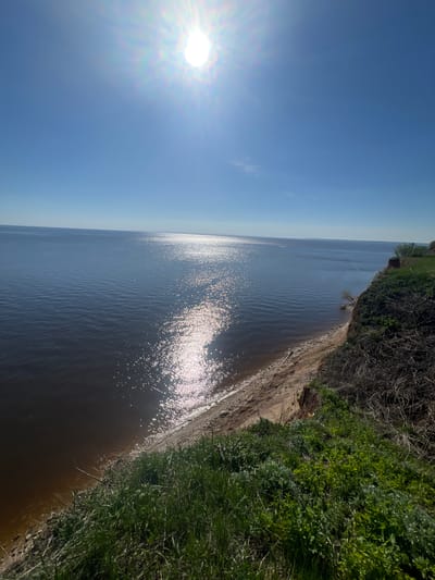 The image depicts a riverbank scene in Шуран, Russia, under a bright, sunny sky. The sun is positioned high in the sky, casting a strong glare on the water's surface. The riverbank is lined with green vegetation, and there is evidence of erosion along the edge. The water appears calm. No people or objects other than natural elements are visible.