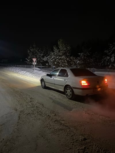 A silver car is positioned on a snowy roadside at night. The car's taillights are illuminated and a haze surrounds its rear. Tire tracks are visible on the snowy ground. A snow-covered tree stands beside a road sign. The scene is dimly lit by the car's headlights and other unknown source. The sky appears dark and clear.