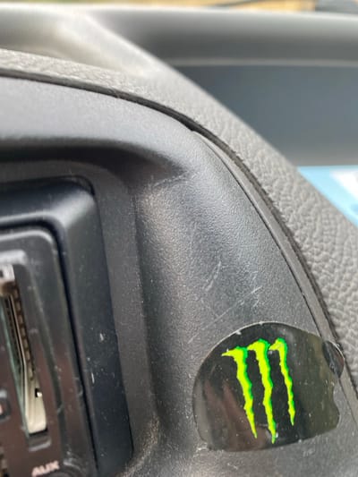 This image depicts the interior of a vehicle, specifically a close-up view of the dashboard area. The dashboard is constructed from textured black plastic, exhibiting visible dust accumulation and minor surface scratches. A portion of a car stereo unit, also black, is visible on the left side, featuring a horizontal slot and a partially obscured "AUX" label at the bottom. Affixed to the lower right section of the dashboard is a black oval-shaped sticker displaying the iconic three-pronged, neon-green "Monster Energy" claw logo. The sticker shows slight wear and edge peeling. The background is out of focus, indicating a view through a windshield or window. No people or discernible actions are present. The precise location in the city of La Tour-d'Aigues, France, cannot be determined from the visual information provided within the image.