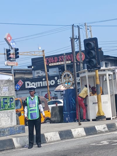 In Animashawun, Nigeria, a traffic officer stands on a street, directing traffic. The officer is wearing a dark uniform with a bright green and blue vest that says "POLICE." Behind the officer, a yellow auto-rickshaw is parked. In the background, there are several businesses, including a Domino's Pizza and a Cold Stone Creamery. Traffic lights are visible, and power lines crisscross the sky. A white booth with the words "TRAFFIC OFFICER" is also present, with two individuals inside.