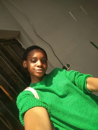 A singular young adult female is depicted in a close-up, upward-angled self-portrait. She possesses dark braided hair, a dark circular earring in her left ear, and a melanin-rich skin tone exhibiting highlights from direct sunlight on her forehead, nose, and cheeks. She wears a bright green, textured, long-sleeved sweater. A light-colored strap is visible on her right shoulder. Her right arm, bent at the elbow, occupies the lower right foreground. The background features a dark, vertically-striped structure on the left, possibly a door or window, with strong linear shadows. The upper and right sections show a light grey wall or ceiling with a thin, dark electrical cable running diagonally. Two pale, rectangular fixtures with dark centers are visible on the mid-right wall, above which a short green rod or pipe segment is partially visible in the upper right corner. The scene is illuminated by strong, direct sunlight casting prominent highlights and shadows. The female subject is directly facing the camera, implying a self-photographic capture. The scene is located in the city of Cotonou, Benin.
