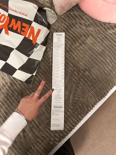 A receipt is laid out on a textured brown surface, positioned slightly to the right of a New Yorker shopping bag. The receipt features several lines of text and numbers, including item descriptions and prices. A hand with painted nails is visible in the lower left portion of the image, with two fingers extended in a peace sign gesture. The shopping bag displays the New Yorker logo in large orange letters against a black and white checkered pattern, with a website address printed at the top. The scene appears to be indoors, with a soft, pink object visible in the upper right corner. The receipt contains information indicating it is from the New Yorker store in Bruchköbel, Germany, with the transaction date of 10.08.2020.