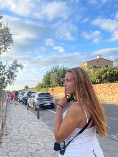 An outdoor scene in Deià, Spain, depicts a paved road and an adjacent stone-patterned sidewalk under a partly cloudy blue sky. In the foreground, positioned right of center, a young adult female with long, light brown hair faces left in profile. She wears a white tank top, holds black sunglasses in her right hand, and her right index finger rests on her chin. A camera with a black strap hangs on her left side. To her left, on the sidewalk, a group of three to four individuals, one in a pink garment, walk away from the viewer. A line of cars is parked along the right side of the road, with a silver-grey Peugeot 208 (license plate 5303 MZM) visible as the last in the row. Dark bollards separate the sidewalk from the parked vehicles. A low stone wall borders the sidewalk on the left, while a tall, dry-stacked stone wall borders the road on the right. A triangular warning sign is affixed to the right stone wall. Beyond the wall and dense green foliage, a traditional stone house with a tiled roof, a chimney, and green-shuttered windows is visible. Olive trees and other vegetation are present on both sides of the road.