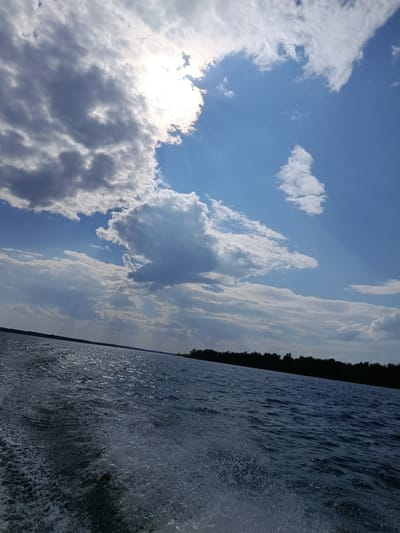 The image shows a wide shot of a body of water with a distinct wake in the foreground, indicating movement, likely from a boat. The water is dark and choppy, reflecting the sky above. A treeline is visible in the distance along the horizon, silhouetted against the bright sky. The sky is a mix of blue and dramatic clouds, with the sun partially obscured by the clouds, casting rays of light. The overall scene suggests a bright, possibly windy day on a large river or lake. The location is identified as Voskresensky District, Russia.