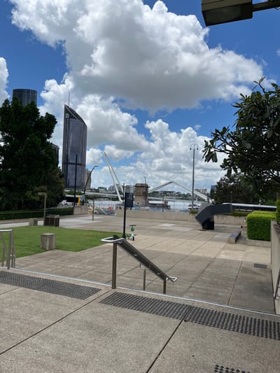The image, originally presented in a vertical orientation but best viewed rotated clockwise, depicts a sunny urban landscape overlooking a river and city skyline. No people are visible, and the scene is static, showcasing public infrastructure and natural elements.

In the foreground and mid-ground on the right, wide concrete pathways are visible, featuring distinct square slabs and tactile paving strips. A metal handrail system runs along a slight incline. Patches of neatly maintained green grass and low concrete structures, possibly benches or planters, are also present. A tall metal pole equipped with sensor-like apparatus, including a green and white element resembling an anemometer, stands in the mid-ground. Further back on the paved area, a dark, angular, sculptural object or piece of street furniture is seen.

The background on the left reveals a wide river with various indistinct structures or small boats. Spanning the river is a prominent white bridge, possibly a cable-stayed or suspension design, with numerous visible white cables and towers. Beyond the river, the urban sprawl includes several modern skyscrapers, one featuring a distinctive angled glass facade. Lush green trees with broad leaves frame parts of the scene in both the foreground and mid-ground.

The time of day appears to be mid-morning to early afternoon, indicated by the bright sunlight and clear shadows cast across the paved areas. The weather is fair, with a vibrant blue sky adorned with large, fluffy white cumulus clouds. The architectural style, prominent bridge, and large river are consistent with a major modern city, aligning with the context of Brisbane, Australia. No legible text is visible on any signs or objects within the image.