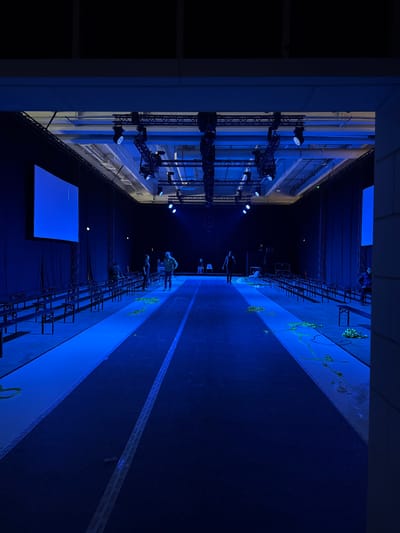 The image depicts an indoor event space in Issy-les-Moulineaux, France, observed from a framed opening on the right. A prominent, dark blue central pathway with a faint lighter stripe runs into the background, flanked by lighter blue strips. Dark, bench-style seating is arranged in multiple rows on both sides of this pathway, facing inward. Overhead, an extensive metal truss system supports numerous stage lights, casting a dominant blue illumination throughout the hall. Walls are uniformly covered in black drapes. Two large, rectangular screens are visible: one on the left displays a solid blue image with minimal white text, and a second, partially obscured screen is on the far right. Seven individuals are present within the space: three are on the left side of the central pathway, two are near the distant end of the pathway in the background, and two are positioned on the right side. Their actions include standing or walking. Scattered clusters of light-colored, possibly yellow or green, organic material resembling foliage are distributed on the lighter strips adjacent to the central pathway, glowing under the blue light. The underlying floor surface beneath the event setup appears light-colored.