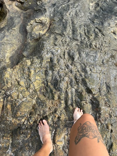 This media file shows a close-up view of a person's feet and a tattooed leg resting on a textured, rocky surface. The rock appears to be wet and weathered, with variations in color from dark gray to golden brown and highlights of white, possibly from salt or sand.
The subjects are a person's right leg and feet, with the feet appearing to have sand or salt residue on them. The leg features a black tattoo with intricate patterns, possibly of a butterfly or a similar winged creature. The scene is outdoors, with the rocky surface suggesting a coastal or natural environment.
The activity is implied to be resting or posing on the rocks. The lighting suggests it is daytime, likely sunny, given the bright highlights on the wet rock. The location cues of a rocky shore and the specific mention of Tambon Phru Nai, Thailand, place this scene in a tropical coastal setting.
Notable details include the rough, uneven texture of the rock, the clear visibility of the tattoo on the person's leg, and the sandy or salty coating on the feet. There is no visible text in the media file. The overall impression is one of casual relaxation in a natural environment.