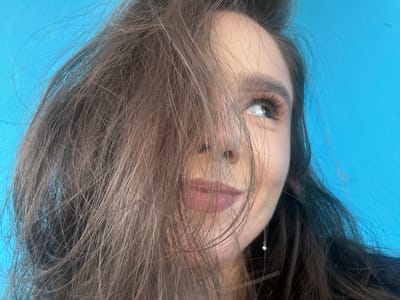 The image is a close-up, medium shot of a young woman's face, taken from a low angle. She has long, brown hair that partially obscures her face, with strands falling over her left eye and across her nose and mouth. Her right eye is visible, looking upwards and to the right, with dark eyelashes and a hint of mascara. Her lips are painted with a soft pink lipstick, and she has a subtle smile. A delicate silver earring with a small pearl or gem is visible hanging from her right ear. The background is a vibrant, solid blue. The lighting suggests it is daytime, possibly outdoors due to the natural light reflecting in her eye, or in a well-lit indoor space. The overall mood conveyed is one of casualness and slight playfulness. There is no visible text in the image. The location context provided is Shulhynka, Ukraine, which does not offer any specific visual cues within the image itself.