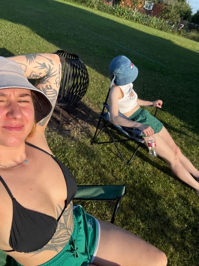 Two individuals are sitting on camping chairs in a grassy backyard. The person in the foreground is wearing a black bikini top and green shorts, with a bucket hat and tattoos on their arm. They are looking towards the camera with their eyes closed. The second person, in the background, is wearing a white tank top and green shorts, with a blue bucket hat. They are sitting facing away from the camera and have a bottle of Coca-Cola on the ground next to them. A black fire pit is visible behind the person in the foreground. The scene appears to be in a rural area, possibly the city of Nemanichy, Belarus, given the presence of a house and trees in the background. The lighting suggests it is late afternoon or early evening.
