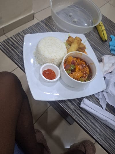 The image depicts an indoor dining scene at Close, Nigeria. A white rectangular plate centrally presents a meal on a striped dark and light grey table surface. The meal consists of a mound of white rice, three golden-brown spring rolls resting on a small portion of shredded vegetables, and a white bowl containing a colorful stew with protein chunks, bell peppers, and onions in a reddish-brown sauce. A smaller white bowl of red dipping sauce accompanies the main plate. To the upper right, a clear plastic container with a translucent lid, a ripe banana, and a light blue plastic pick are visible. A crumpled white paper napkin and a folded white paper napkin are positioned to the right of the plate. The lower portion of a person's dark-skinned legs, partially clad in light blue fabric, is visible on the left and bottom of the frame, with brown fuzzy slippers on their feet, indicating they are seated at the table. The floor beneath the table consists of light-colored tiles.