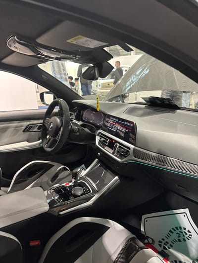This image captures the interior of a modern BMW automobile, likely an M-series, situated within an exhibition or showroom environment in Wolica, Poland. The perspective is from the front passenger seat, looking forward towards the driver's side and through the windshield into the immediate surroundings.

**Vehicle Interior:**
The car features a black Alcantara-wrapped steering wheel with a yellow stripe at the 12 o'clock position and a central BMW logo. The dashboard is equipped with a digital instrument cluster and a wide central infotainment screen, both illuminated and displaying information. Carbon fiber trim is extensively utilized across the dashboard, center console, and parts of the seats. A thin, illuminated teal-colored ambient light strip runs horizontally across the dashboard. The center console includes a gear selector, the iDrive controller, and several buttons, notably a prominent red 'M' button. The sport seats are upholstered in a combination of white and black material, featuring pronounced bolsters and additional carbon fiber accents. The headliner is dark-colored, with an overhead console and passenger sun visor visible, the latter displaying an airbag warning label. A yellow air freshener hangs from the rearview mirror. On the front passenger floorboard, a white paper mat with a black footprint design is secured by a red clip. Some white papers are placed on the dashboard directly beneath the windshield.

**Exterior Environment (visible through windshield):**
The background reveals an indoor setting, likely a convention hall, characterized by bright artificial lighting. Several individuals are present. A male in a grey long-sleeved shirt and dark pants stands in the mid-ground, facing right. Another person is partially visible to his left. Behind these individuals, a light-colored vehicle, possibly cream or white, is displayed with its hood open, revealing engine bay components and a reflection of the ceiling or another structure on its underside. White display panels or walls constitute the distant background. No direct interactions are discernible among the visible people; they appear to be observing the exhibition.