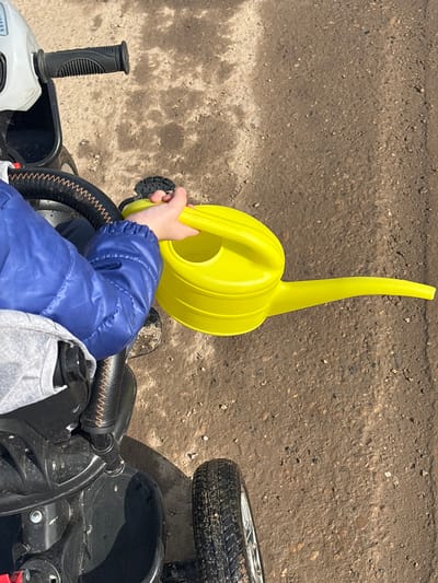 A child is seated on a black tricycle on a dirt and gravel path in Gamovo, Russia. The child, wearing a blue quilted jacket and light grey lower garment, holds a bright yellow plastic watering can in their right hand. The watering can features a single handle and an elongated spout directed towards the right. Visible tricycle components include a black textured handlebar grip on the right, a black seat or armrest with brown stitching, and a black wheel with a silver hub, partially soiled with dirt. The ground consists of compacted light brown soil intermixed with small stones.