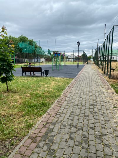 The image depicts a paved walkway in an outdoor public space in Zapadny, Russia. The walkway, constructed of interlocking grey and reddish-brown cobblestones, leads away from the viewer into the distance, bordered on the right by a chain-link fence with green covering. To the left of the walkway, a grassy area is visible, with a young tree in the foreground and a wooden bench with a metal trash receptacle. A person, appearing to be a young male wearing a grey shirt and dark shorts, is seated on the bench. Beyond the bench, a playground area is visible, featuring colorful structures and green netting. Further in the background, a sports field with a fence and some buildings can be seen under a cloudy sky. A single lamppost stands near the playground.