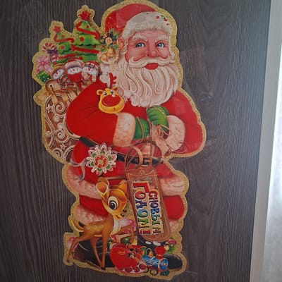 The image displays a flat, festive Christmas decoration of Santa Claus with a sack of gifts. He is depicted with a large white beard and mustache, blue eyes, and rosy cheeks, wearing his iconic red suit trimmed with white fur. To his left, a miniature Christmas tree adorned with ornaments and a red star is visible, along with three snowmen and a wrapped gift. Santa holds green-gloved hands, and his sack appears to be made of burlap with gold glitter accents.
A small reindeer with a yellow face and brown spots stands near Santa's feet, accompanied by a red heart-shaped gift, a toy train, and other festive items. A wooden tag hanging from Santa's sack reads "С НОВЫМ ГОДОМ!", which translates to "Happy New Year!" in Russian. The decoration is affixed to a dark wooden surface, likely a door or wall, characterized by vertical grain patterns. The lighting suggests an indoor environment. The image does not provide any specific details about the city of Bugulma, Russia, or any activities taking place within it.