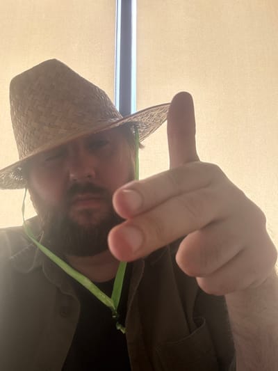 A single adult male is depicted from the chest up, positioned centrally in the frame. He has a dark beard and is wearing a light-colored woven straw hat with a pointed crown and wide brim, tilted low over his forehead, obscuring his left eye. His visible right eye looks directly towards the viewer with a neutral to serious expression. He wears a dark olive-green collared shirt, open at the top, revealing a dark undershirt. Around his neck hangs a bright lime-green lanyard with white text, which includes "ЗЕЛЕНОГОРСКОЕ" and a partial web address ending in ".ru". His right hand is raised towards the viewer, with his index finger pointed upwards and his middle finger extended forward, forming a gesture resembling a handgun. The background is uniformly light beige or off-white, possibly a wall or a window with closed blinds, with a thin vertical dark strip visible on the far left. The scene is located in Зеленогорское сельское поселение, Ukraine.