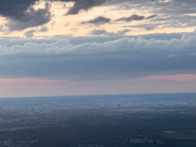 The image appears to be an inverted view from above, likely taken from an aircraft, depicting an evening or night scene. The top portion shows a darkened landscape with sparse lighting, indicative of a city or urban area at night. The presence of darker patches suggests fields or less densely populated zones. A distinct line separates this urban area from a layer of atmospheric haze or smog below, which transitions into a pinkish-purple hue. Below the haze, various layers of clouds are visible, ranging from dark gray to lighter, white-tinged formations, suggesting a low-lying cloud cover during sunset or twilight. The city is Schönefeld, Germany.