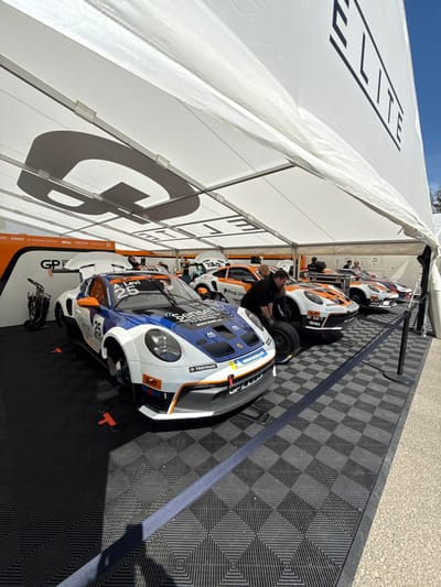 A white tent with "ELITE" printed on the side is pitched over a black and white checkered floor. The tent is set up over a pit lane at the 24 Hours of Spa race track. The tent's canvas is partially collapsed, revealing a clear blue sky. A white Porsche race car with blue and orange accents is parked in the center of the frame. It is the only vehicle fully inside the tent. The car has the number "25" on its side and "A. Levi" on the windshield. It has the words "Sensei" and "Mobil" on its side. A man wearing a white shirt, dark shorts, and black shoes is working on the tire of the Porsche race car. There is another white Porsche race car with blue and orange accents parked to the right of the Porsche. The second Porsche has the number "18" on the side and the words "Prop-Tech" and "Gassan" on its side. There is a third white Porsche race car with blue and orange accents parked further to the right. The third Porsche has the number "17" on the side. Several people in the background are standing near other cars, and a few are walking in the area. The pit lane is located in the town of Stavelot, Belgium, at the coordinates 50.441° N, 5.969° E.