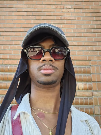 A man is photographed in front of a brick wall. He is wearing sunglasses, a headscarf, and a light-colored shirt with a red belt. There is a lipstick mark on his neck. The photo was taken vertically and is rotated. No location in Massy, France can be inferred from this image.